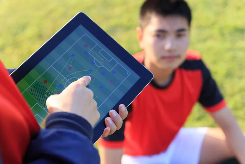 Treinador mostra esquema tático no tablet a jogador de futebol, base da análise de dados nas apostas esportivas