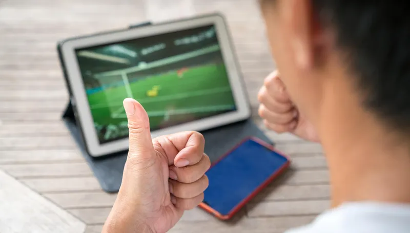 Torcedor faz sinal de positivo diante de tablet com jogo de futebol e celular, apostas online