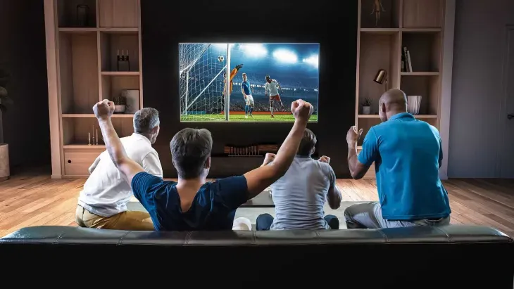 Quatro amigos no sofá comemoram um gol exibido na TV da sala ao assistir TV online free